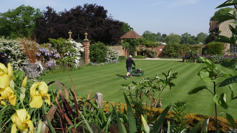 A garden mows the lawn in the formal garden at Packwood, Warwickshire. Spring flowers can be seen bordering the lawn.